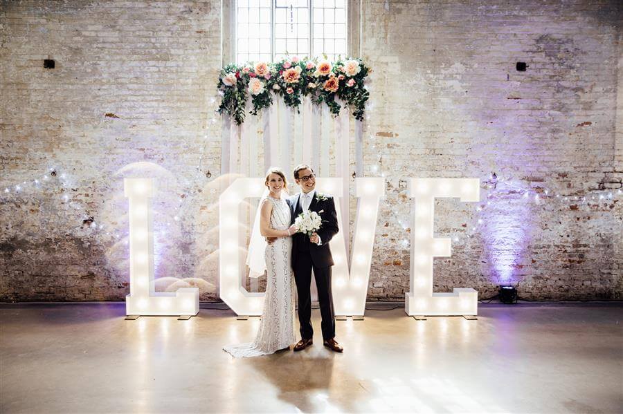 Happy couple stood in front of light up LOVE letters against white painted wall with floral archway