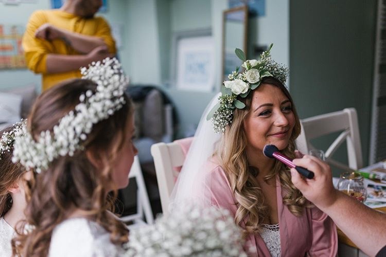 Bride having makeup applied smiling off screen as small girl looks on wearing a flower crown