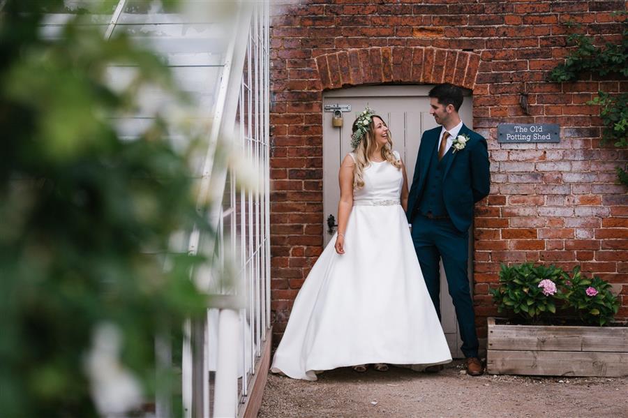 Bride and groom smile at each other in a garden in front of red brick wall by Emily & Katy Photography