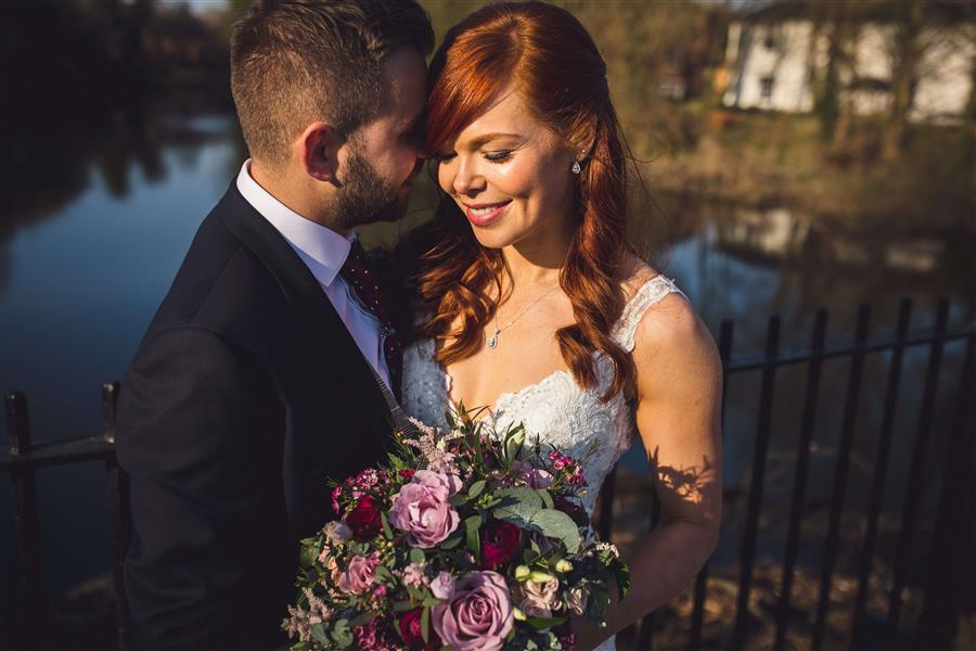 Wedding couple portrait of smiling bride with red hair and pink bouquet in front of a river