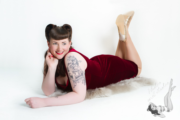 Lady lying on her front wearing red velvet dress with vintage hair and makeup, and large arm tattoo