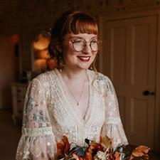 Red haired bride with fringe and glasses wearing lace wedding dress and round glasses smiles out the window