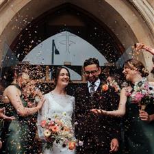 Grinning couple hold hands outside of church as guests throw confetti around them