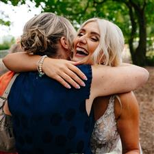 Blonde haired bride hugs a wedding guest and laughs, she has a french manicure, pink lipstick and looks joyful