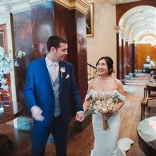 Groom in blue suit and bride in white off the shoulder wedding dress look at each other walking down hall way, bride has dried flower bouquet