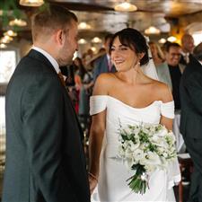 Tanned brunette bride with white lily bouquet smiles at her groom at the end of the aisle