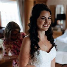 Brunette bride with long hair smiles off camera as her Mum ties up her wedding dress