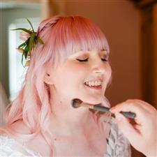 Pink haired bride with winged eyeliner smiles as powder is applied