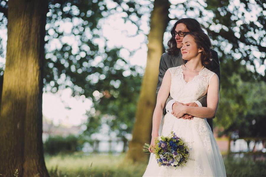 Husband and wife embrace near trees with mottled sunlight