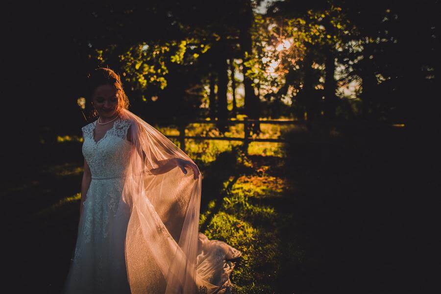 Bride plays with her veil in a field with mottled warm sunlight by Kathryn Edwards Photography
