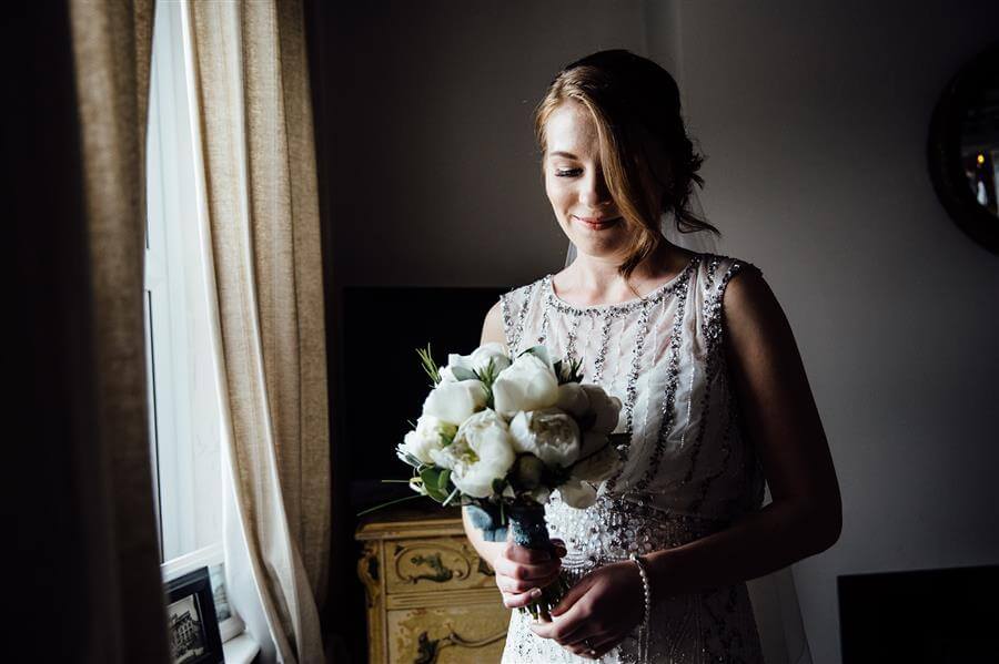 Bride wearing white elegant gown looking down at her white bouquet in a window