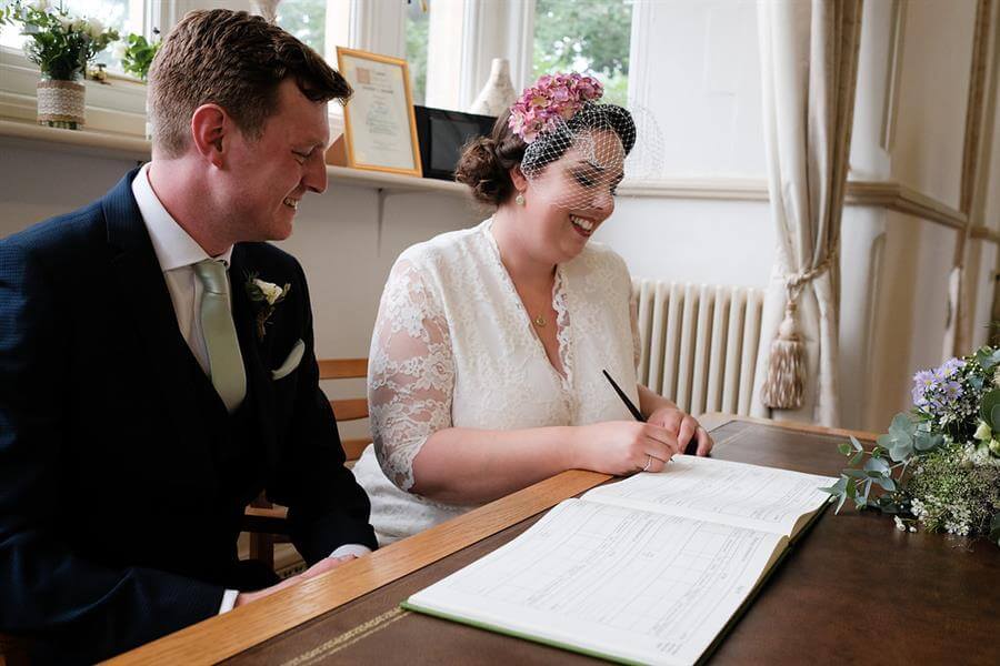Bride wearing white birdcage vintage veil signing the register with her husband
