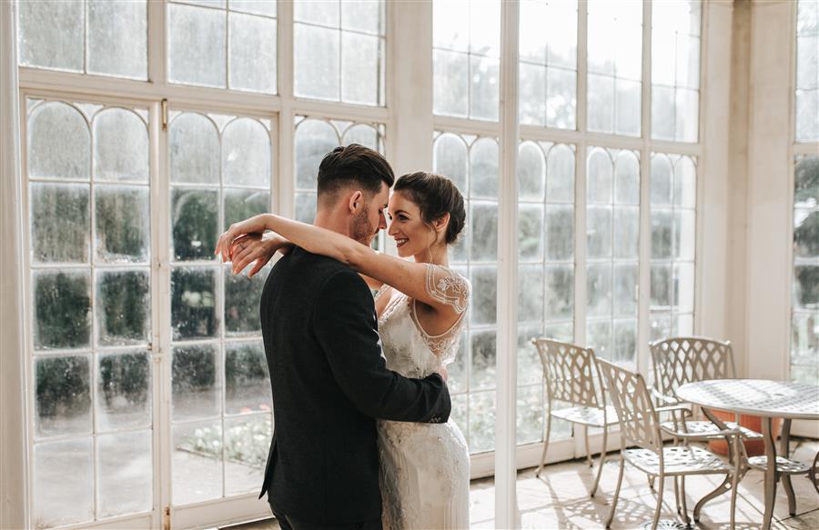 Bride and groom embrace in front of large windows