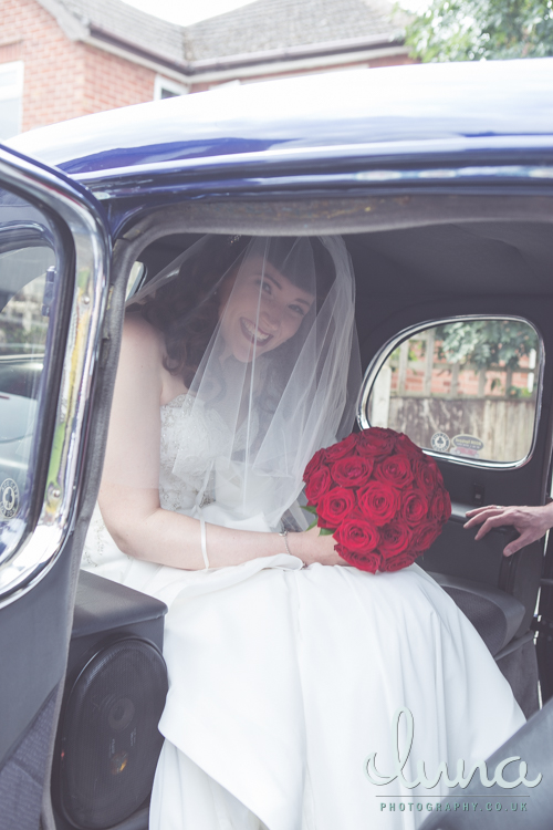 Smiling bride with white veil holds a bouquet of red roses while sitting in a hot rod